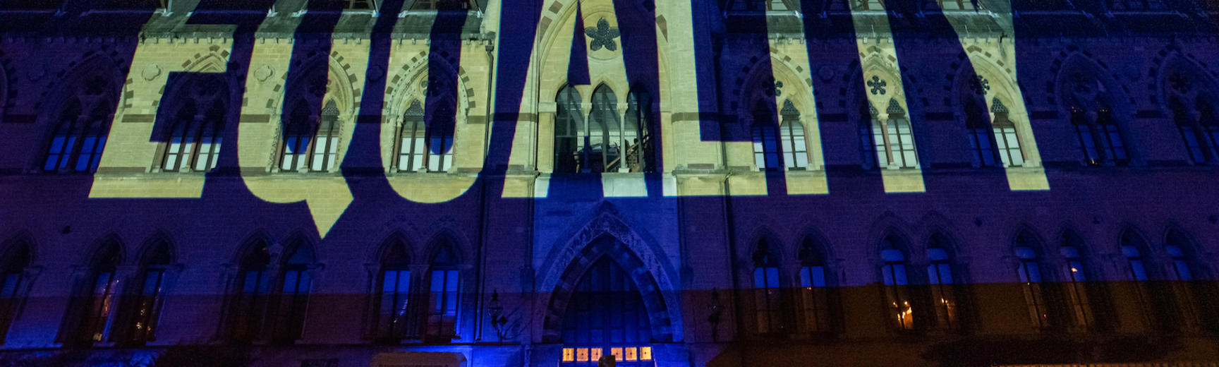 The word EQUALITY projected in yellow light onto the facade of the Museum at night. 