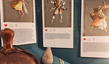 Close up view of a display case showing objects including a model boat, fish hooks and a container made from a large gourd, with paintings of humanoid-insect figures hanging above