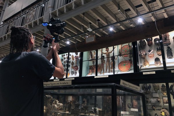 a person lifts a digital camera on a gimble above their shoulders, pointed at objects display in tall glass cases at the museum.